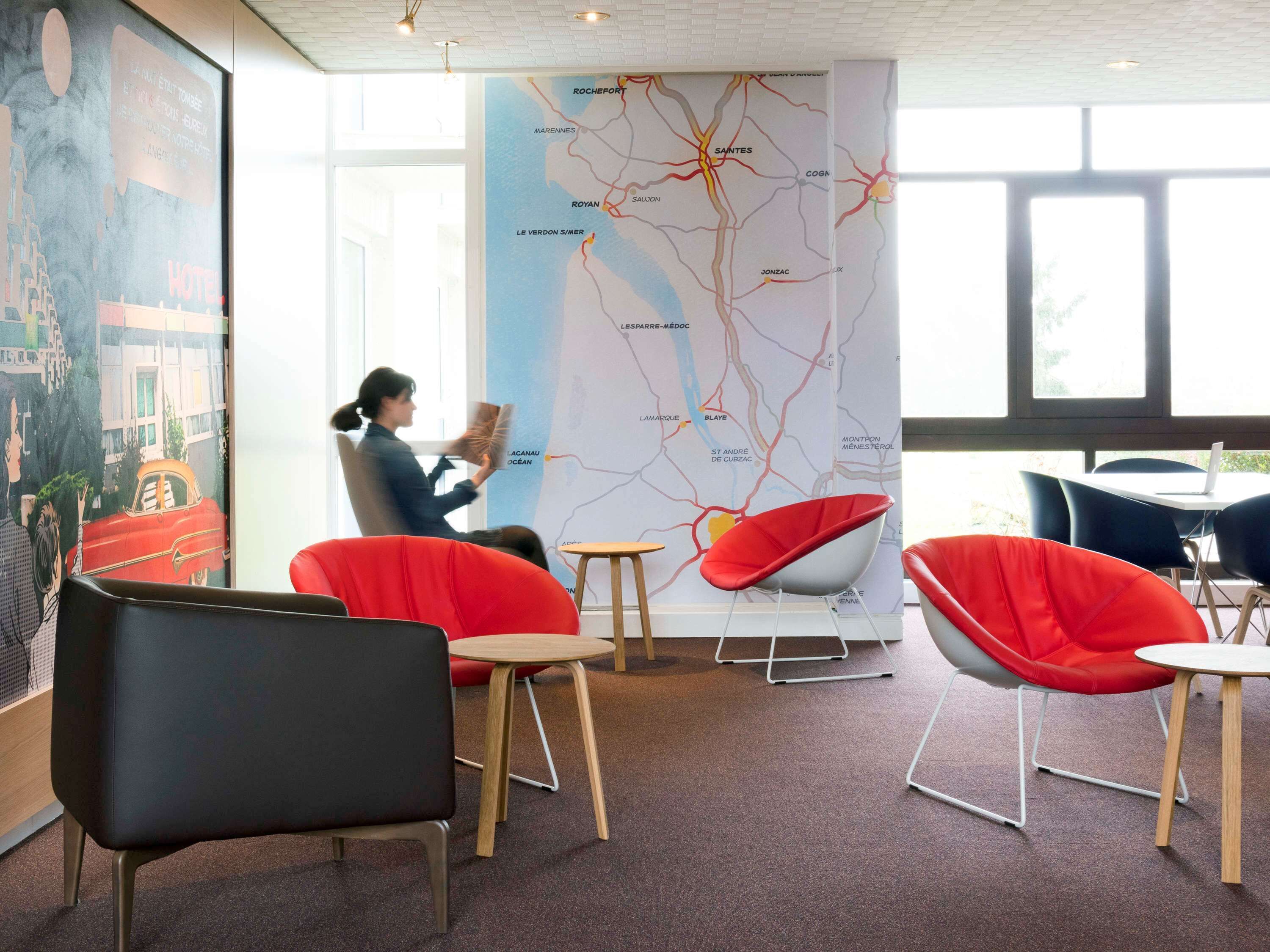 A modern hotel lobby with red and black chairs, a large map on the wall, and a person reading a book.