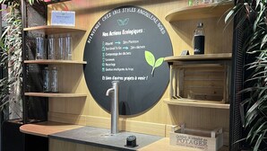 A wooden counter with a black circular sign detailing ecological actions, surrounded by shelves with glass jars and a water bottle. - ibis Styles Angouleme Nord (Champniers)