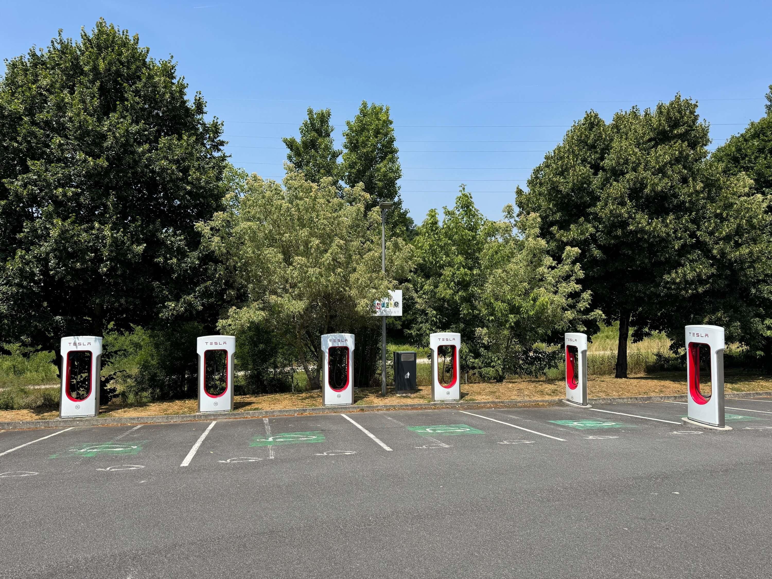 A parking area with four Tesla charging stations, marked parking spaces, and a sign indicating a nearby park.