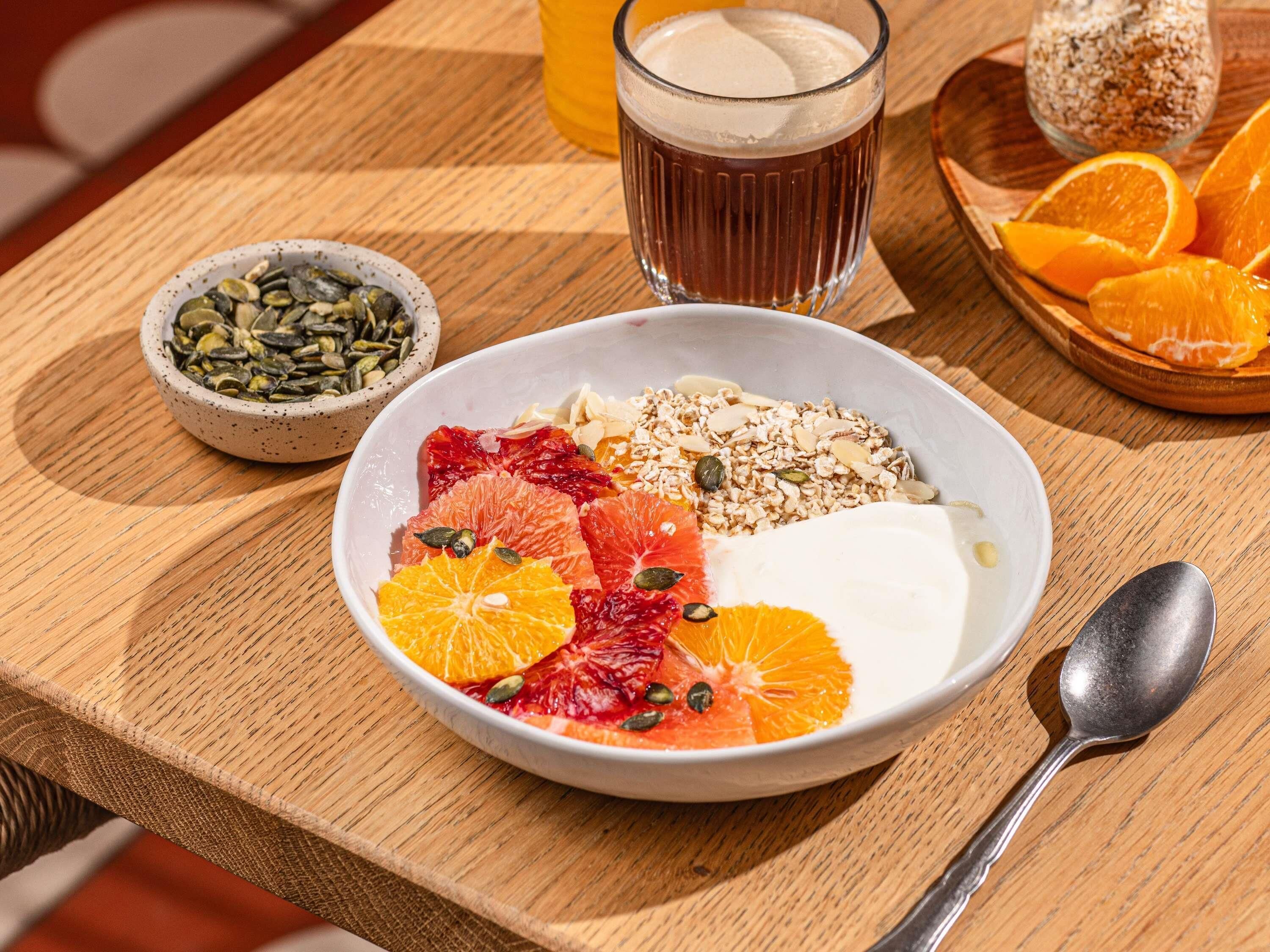 Een ontbijtsetting met een kom yoghurt belegd met fruit en granola, een glas sinaasappelsap, en een schaaltje zaden.