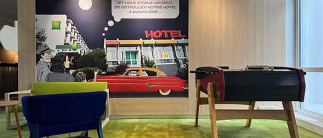 Una habitación con un mural de un hotel antiguo, alfombra verde, una silla azul y una maleta roja sobre un atril.