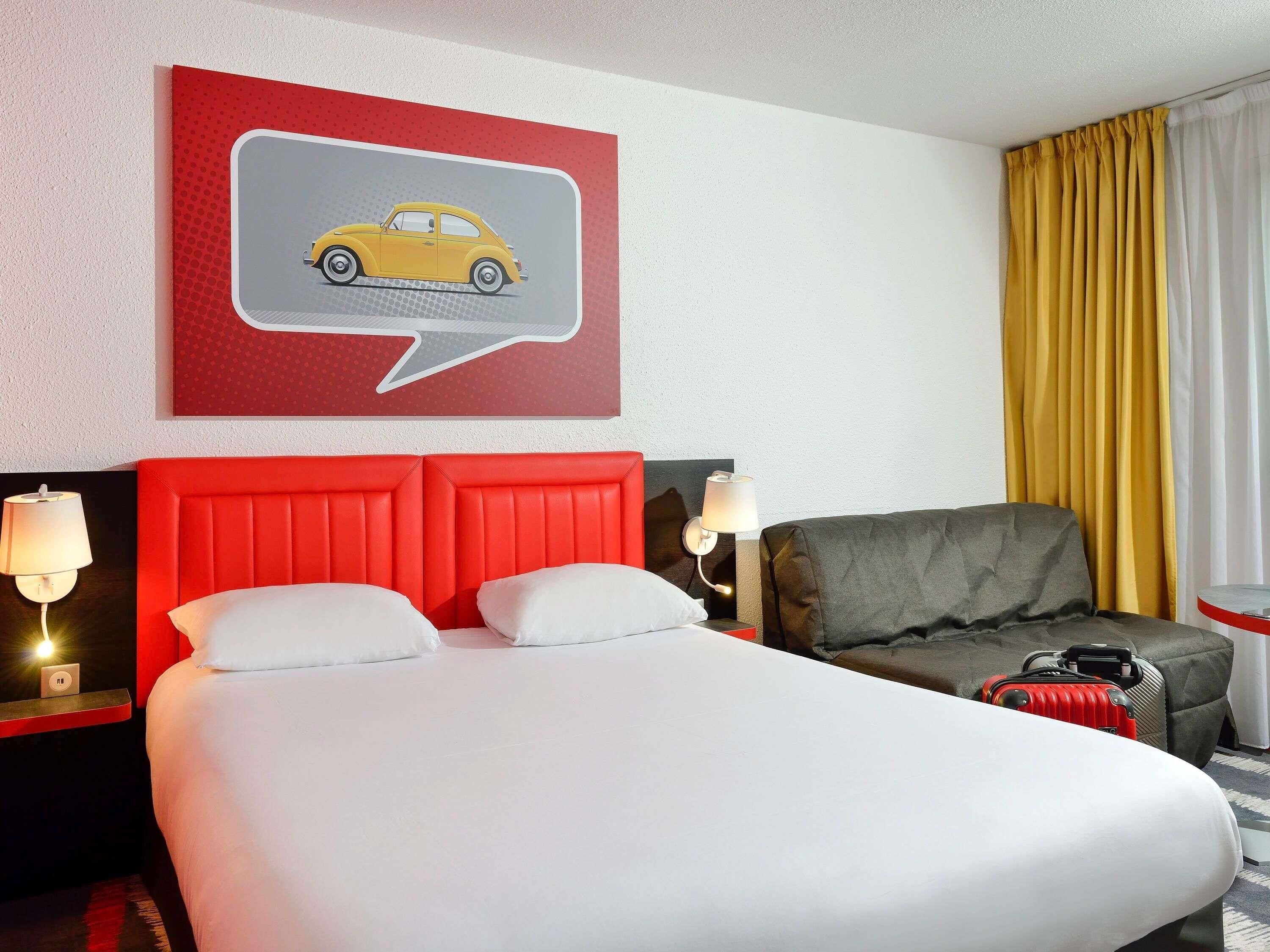 Una habitación de hotel con una cama, un cabecero rojo, un sillón gris, una ilustración de un coche amarillo en la pared y una ventana con cortinas amarillas.