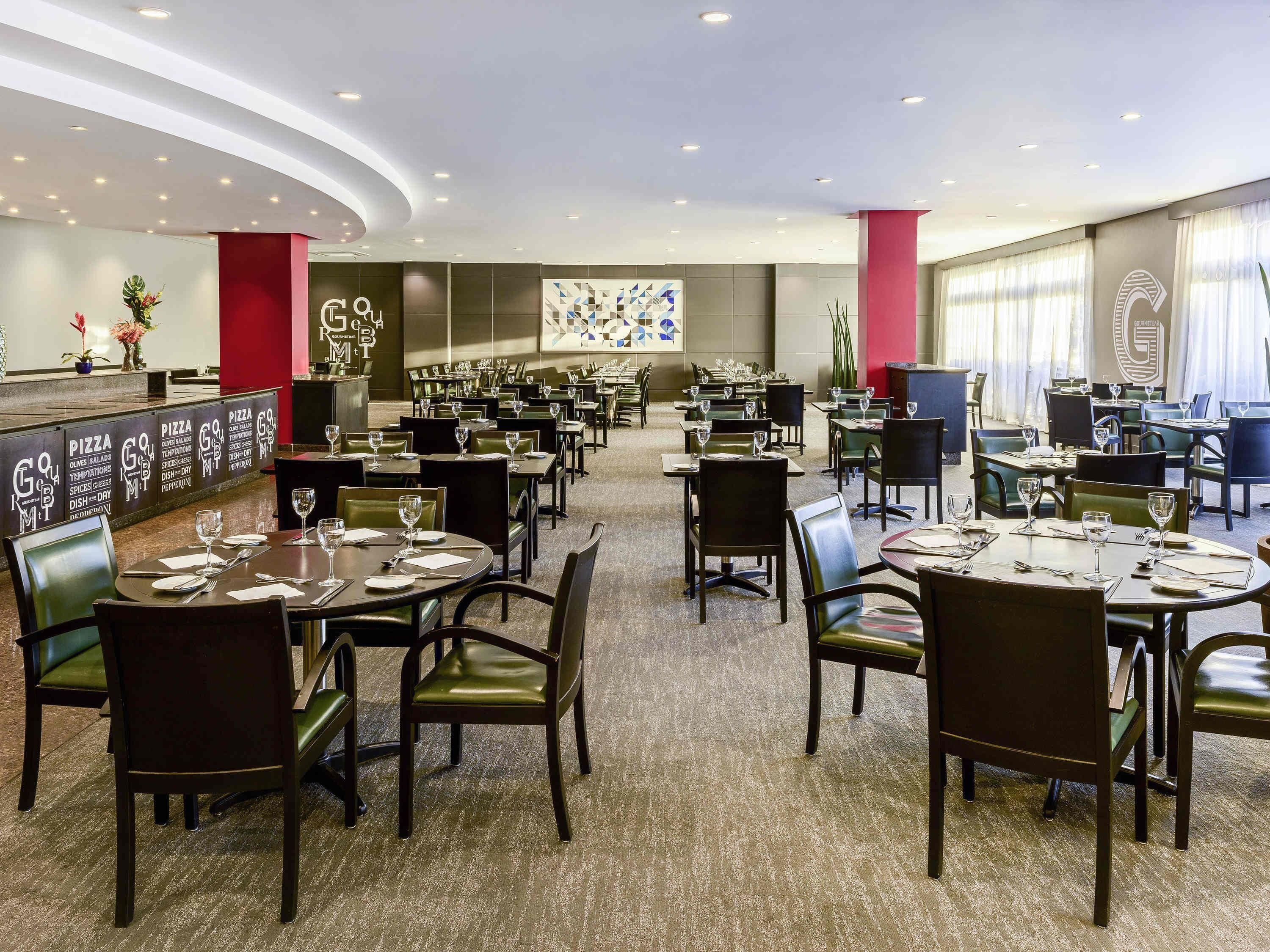Um restaurante amplo, com várias mesas e cadeiras, um bar e uma parede grande com um padrão geométrico.