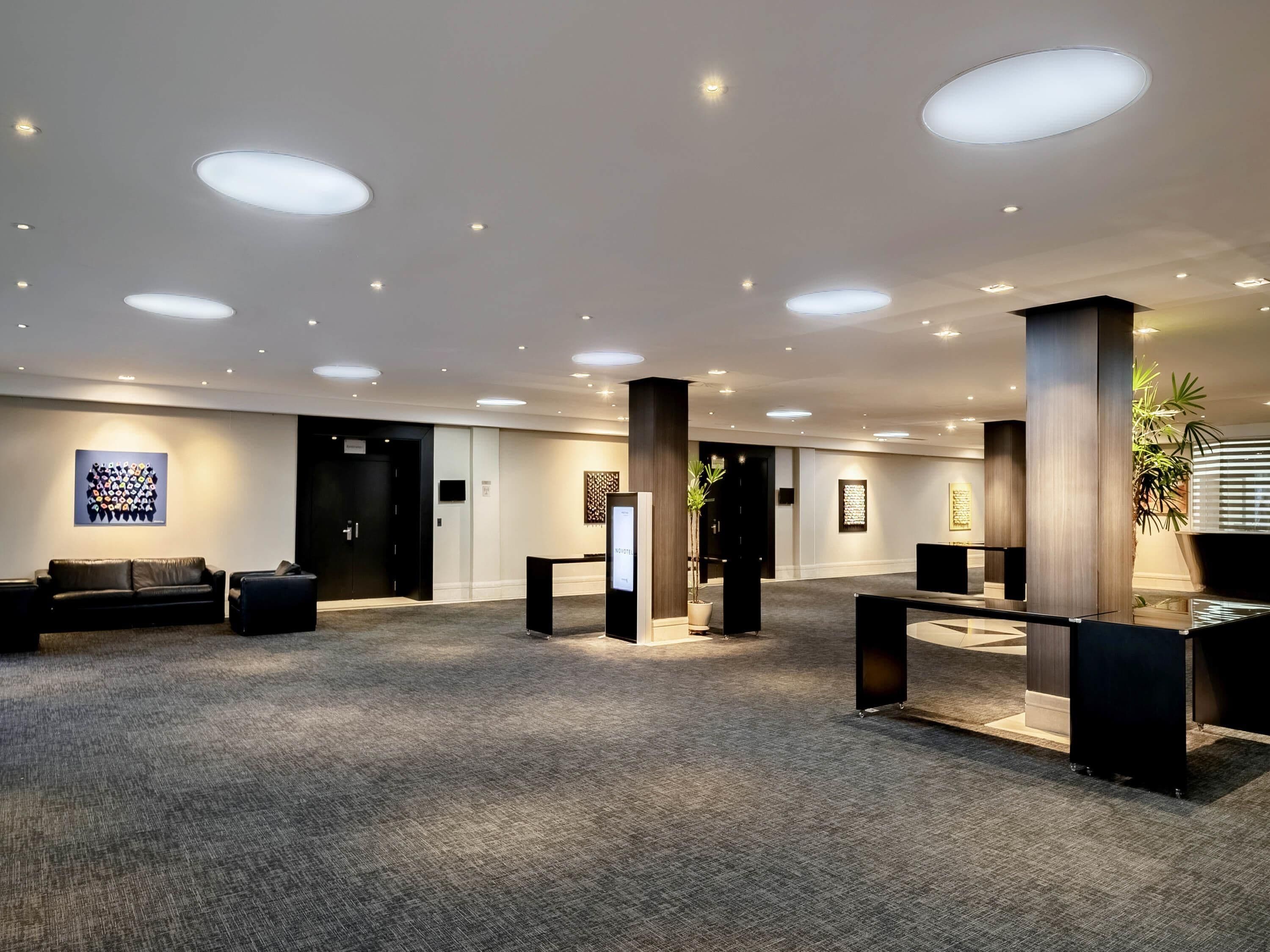 Un hall d’entrée spacieux, doté d’un tapis gris, de canapés noirs, d’un comptoir de réception et d’une grande plante en pot.