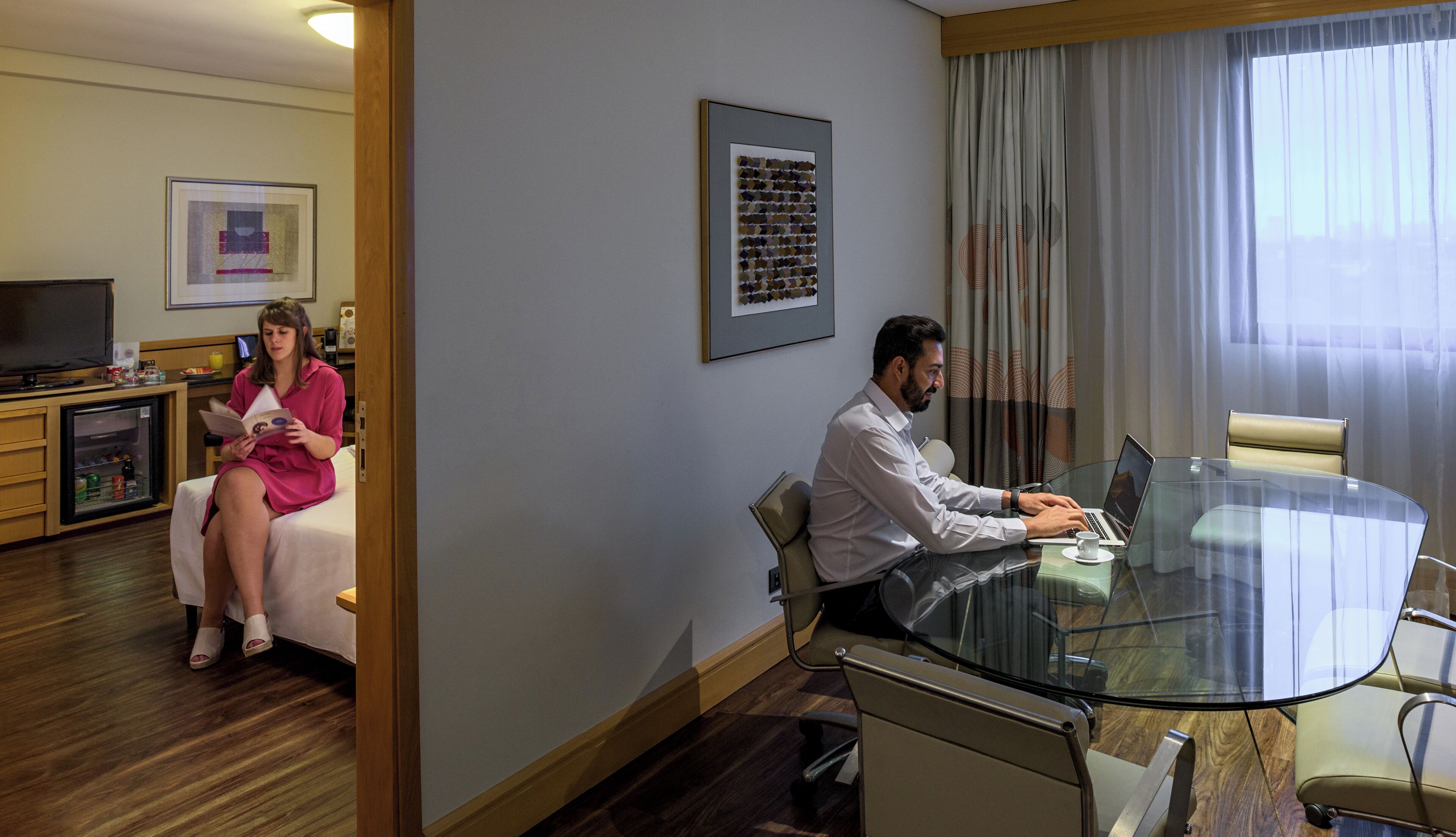 Quarto de hotel com mesa de vidro, televisão, frigorífico e cama.
