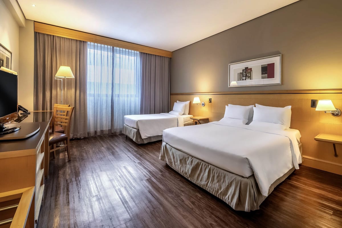 Quarto de hotel com duas camas, uma mesa com computador, uma cadeira e janela com cortinas.