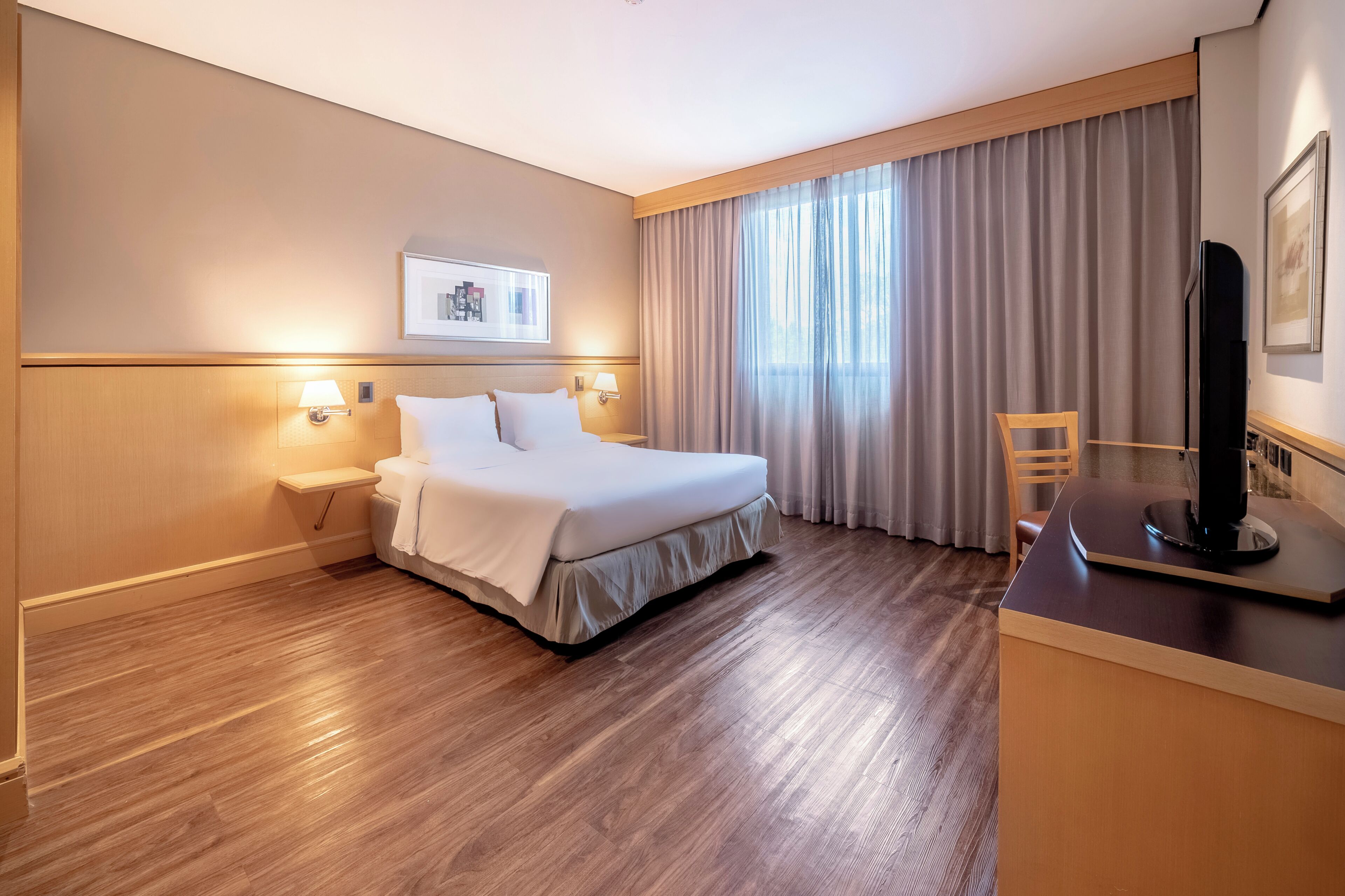 Una habitación de hotel con cama, lámparas de noche, un escritorio con televisor de pantalla plana, una silla y un ventanal con cortinas.