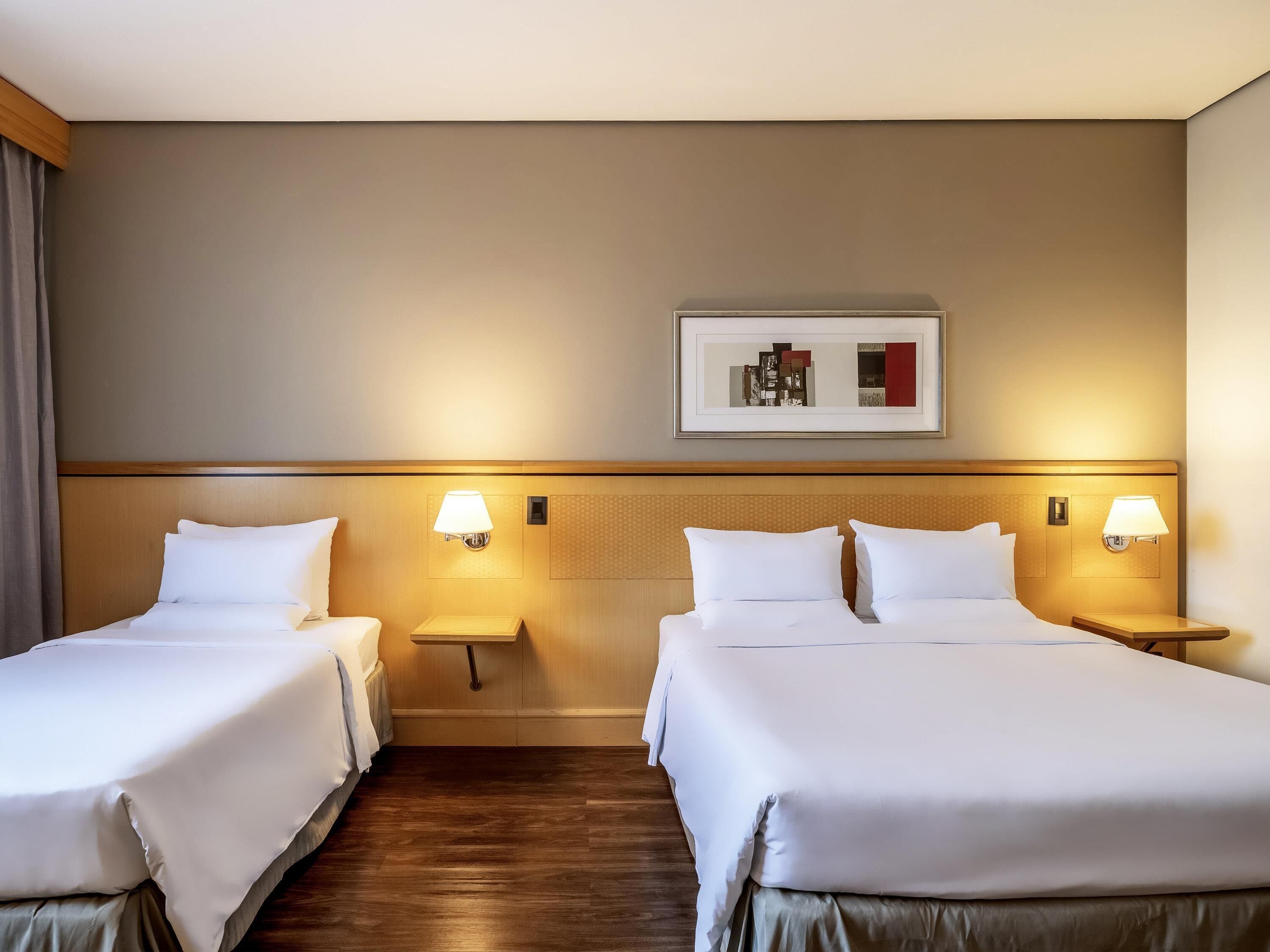 A hotel room with two beds, wooden headboards, bedside tables with lamps, a framed artwork on the wall, and a wooden floor.