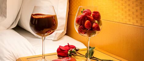 A bed with white linens, a bedside table with a glass of red wine, a glass of dessert with strawberries, a red rose, and a heart-shaped box.