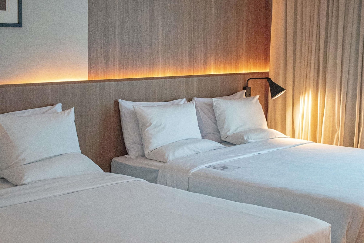Quarto de hotel com duas camas, cabeceira, luminária de cabeceira e cortina.