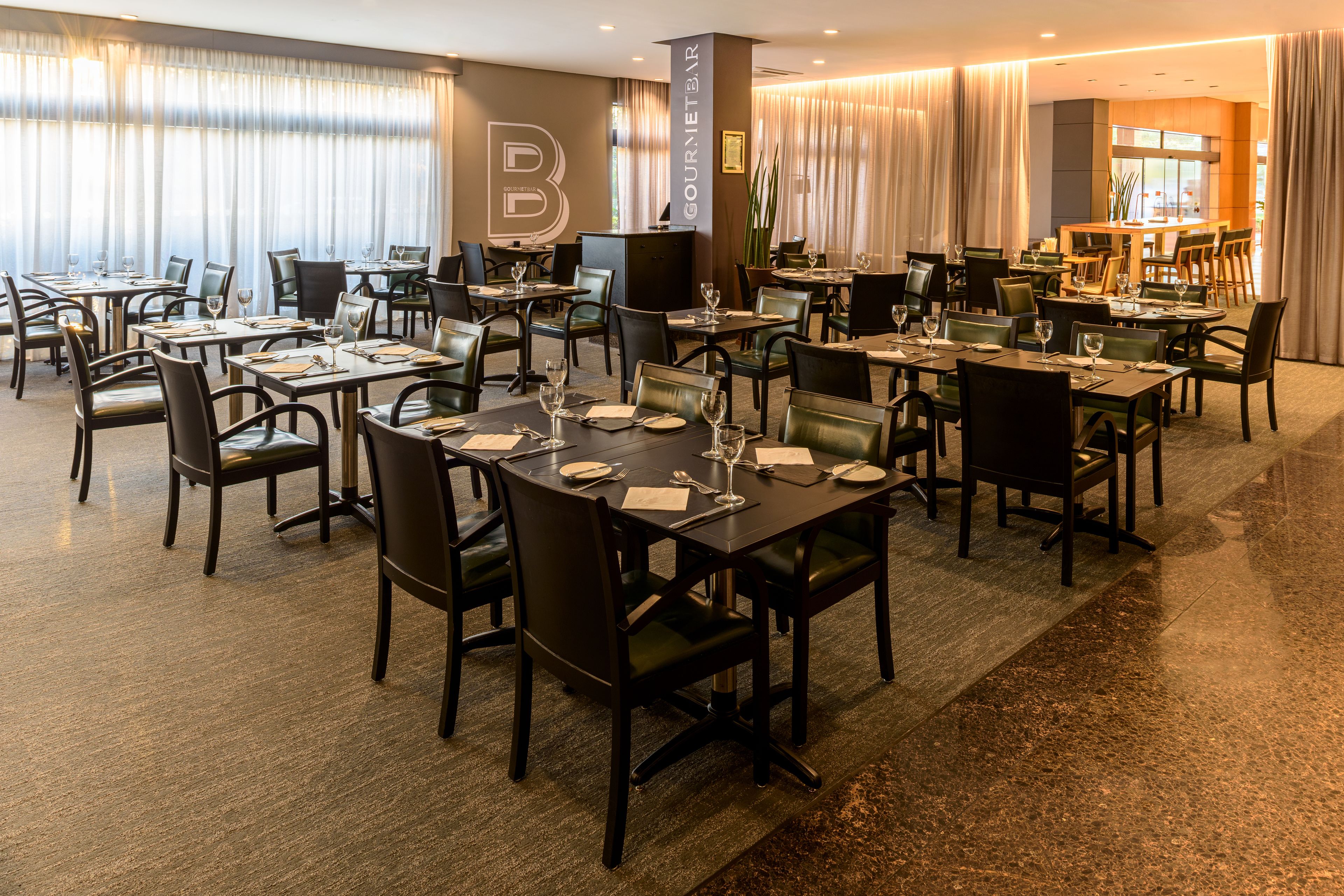 A hotel dining area with multiple tables and chairs, a large sign reading "GOURMET BAR", and a view of the kitchen in the background.
