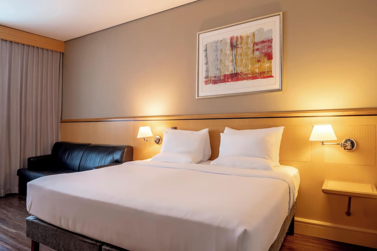 Quarto de hotel com uma cama grande, duas luminárias de cabeceira, uma poltrona de couro preto, uma pintura abstrata emoldurada e uma cabeceira de madeira.