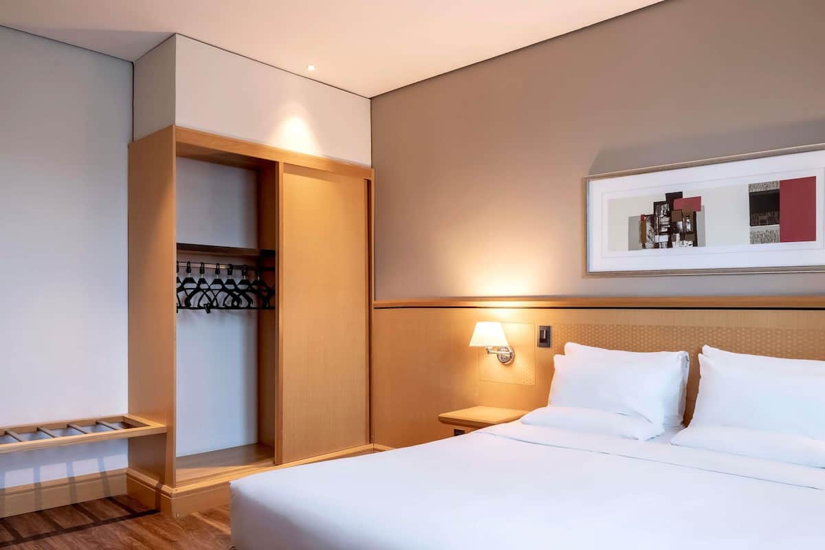 Quarto de hotel com cama, mesa de cabeceira, luminária na parede, porta-casacos e um quadro emoldurado.