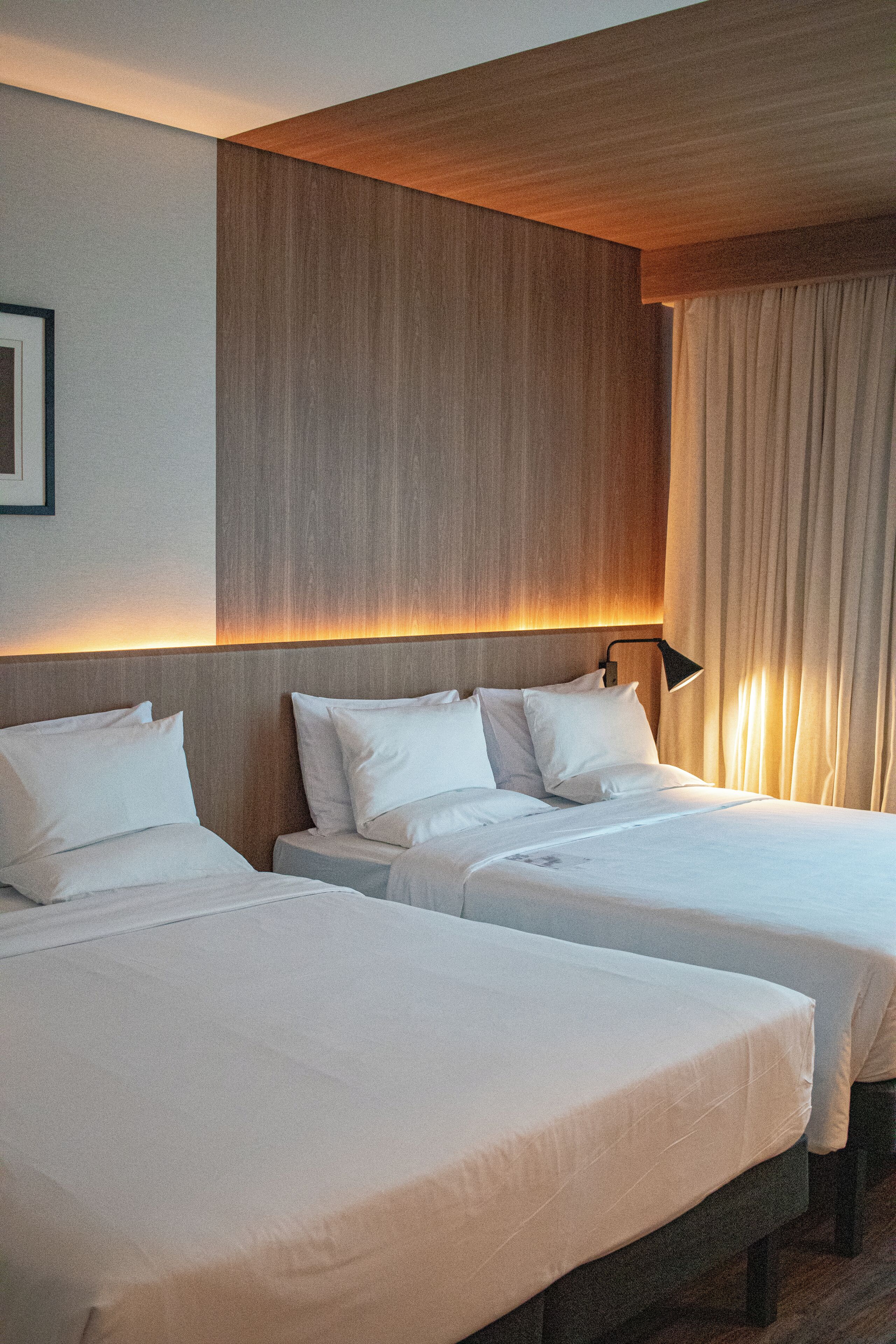 Ein Hotelzimmer mit zwei Einzelbetten, einem Holzkopfteil, einem gerahmten Bild an der Wand und einem Vorhangfenster.