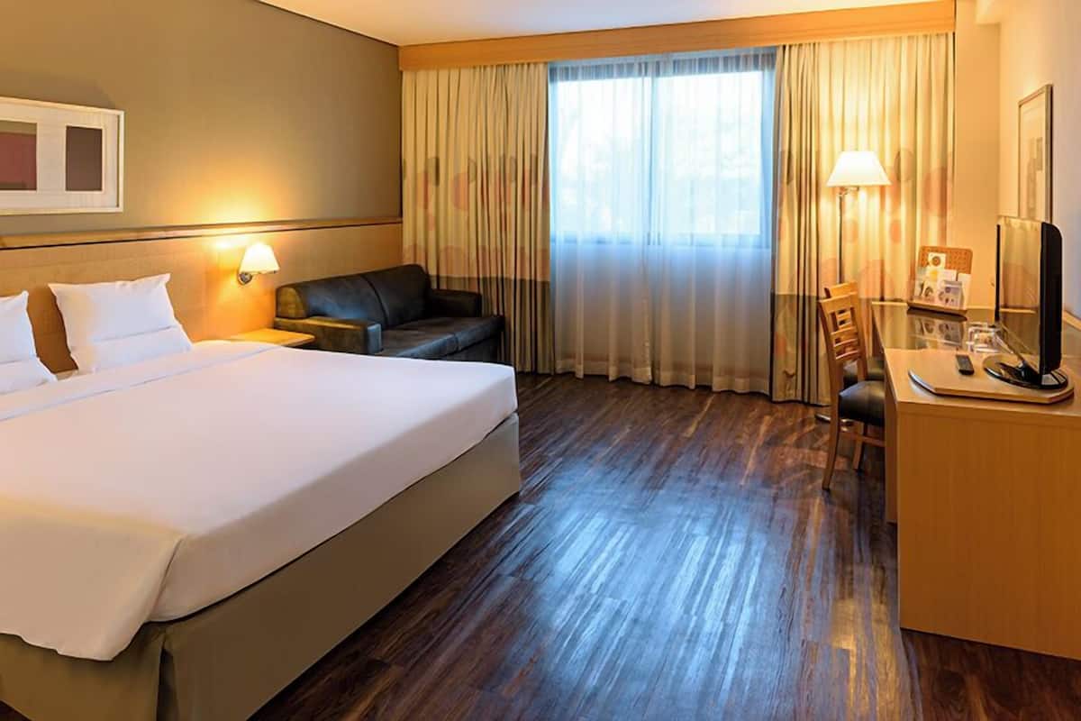 Quarto de hotel com cama grande, escrivaninha, cadeira, luminária e televisão.