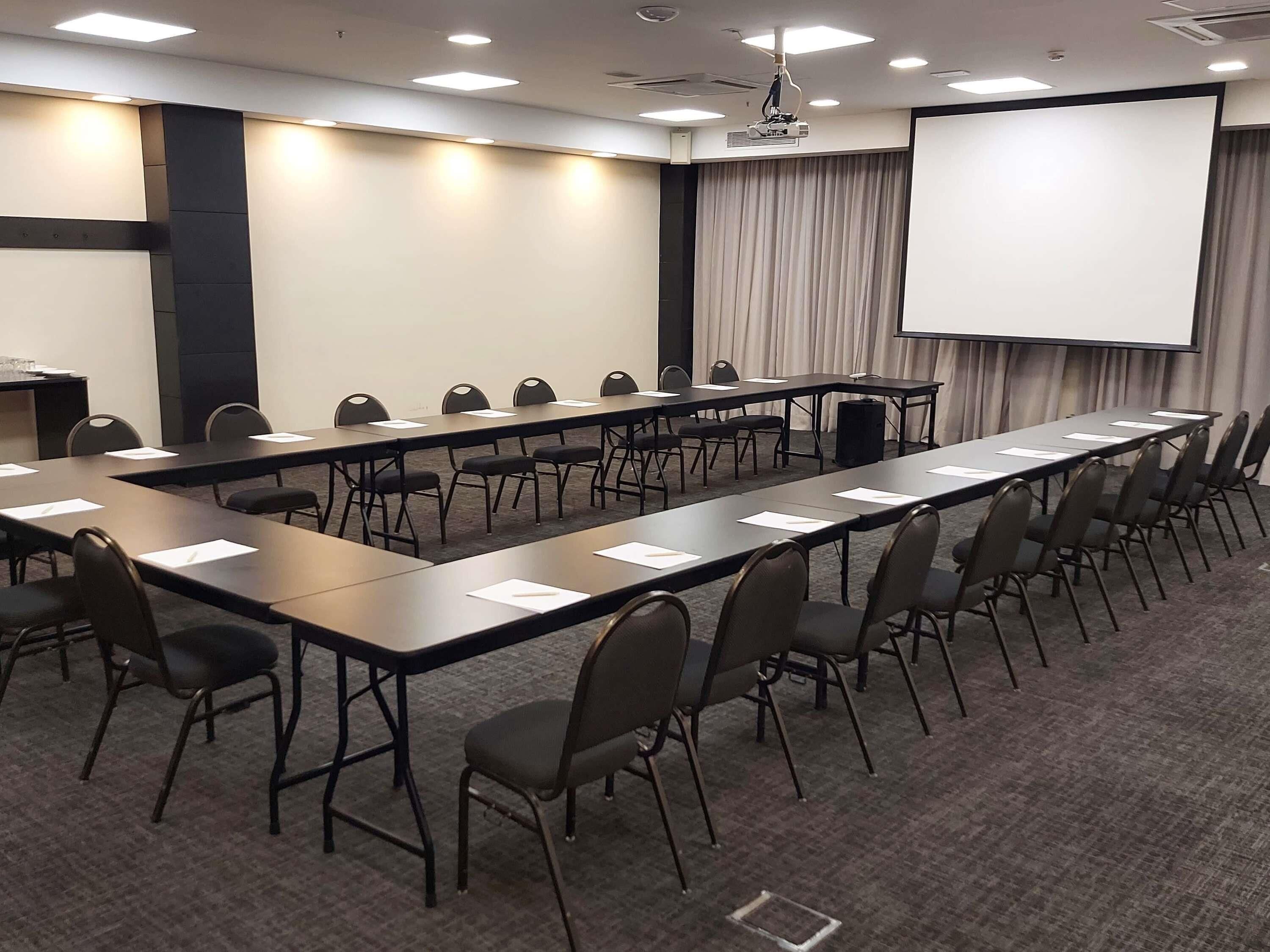 Un salón de conferencias con mesas largas, sillas, un proyector y una pantalla.
