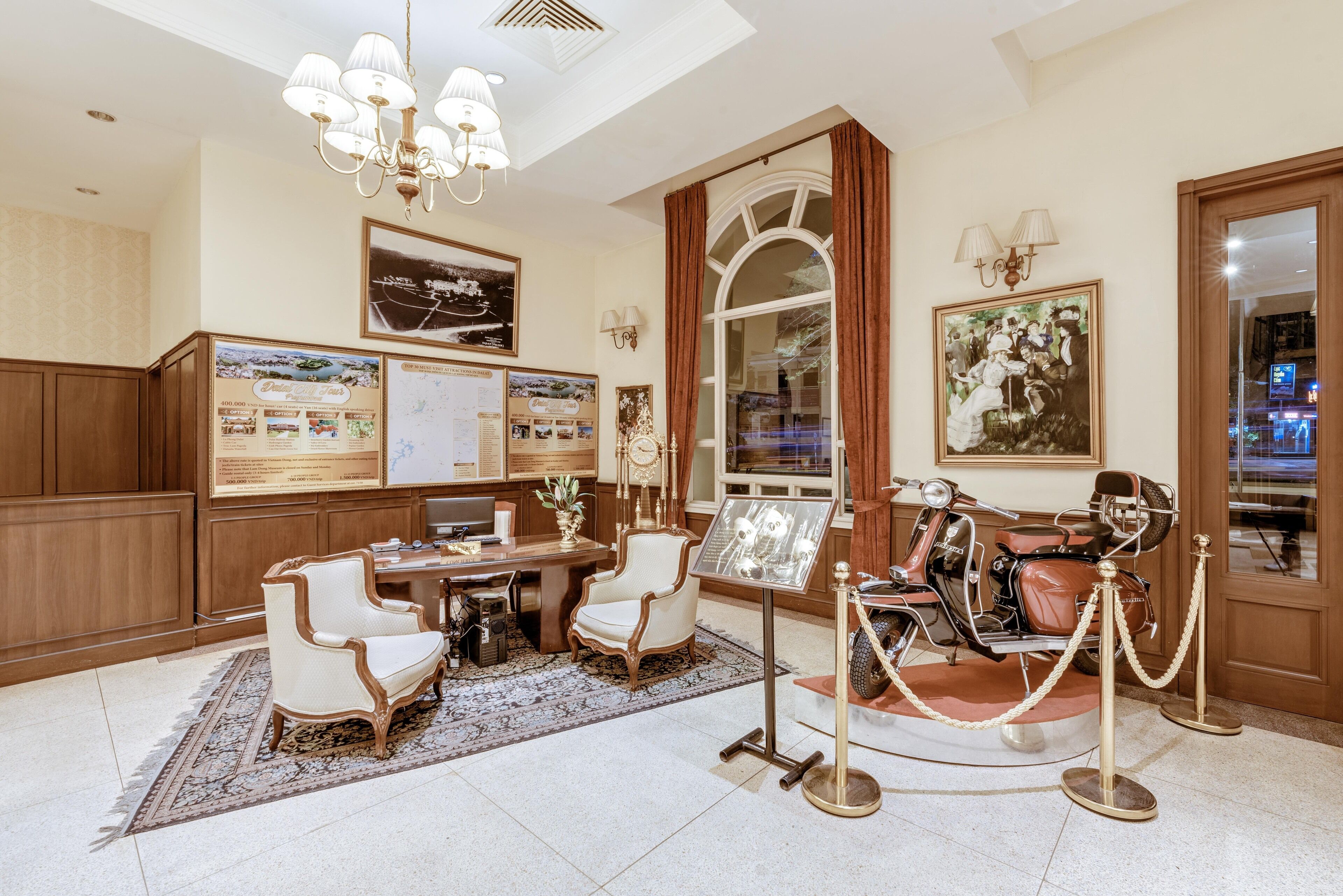 A hotel lobby with a classic design, featuring a wooden reception desk, a seating area with armchairs, a vintage motorcycle on display, and a chandelier.