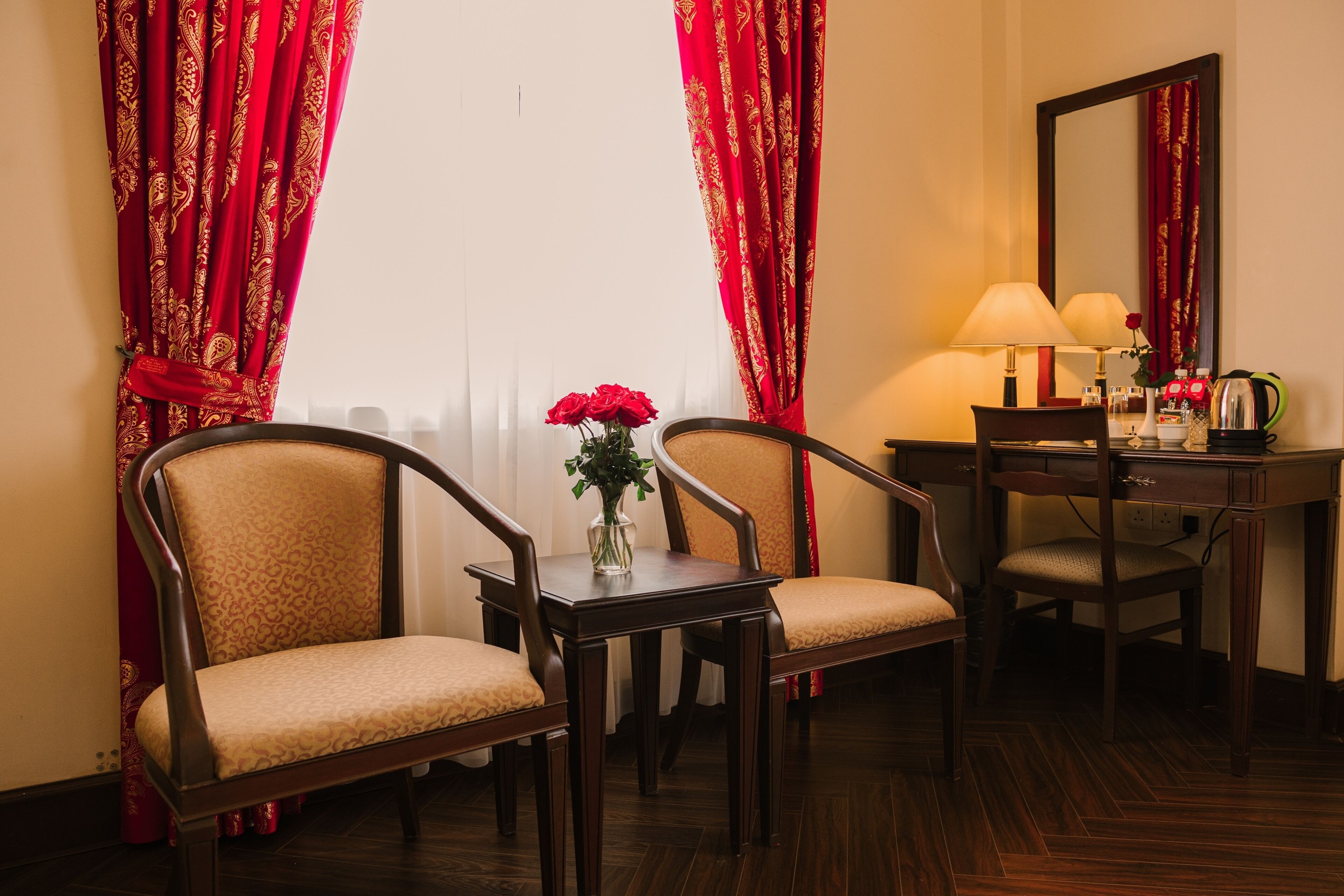 A hotel room with two armchairs, a small table with a vase of red flowers, a desk with a lamp, and a mirror.