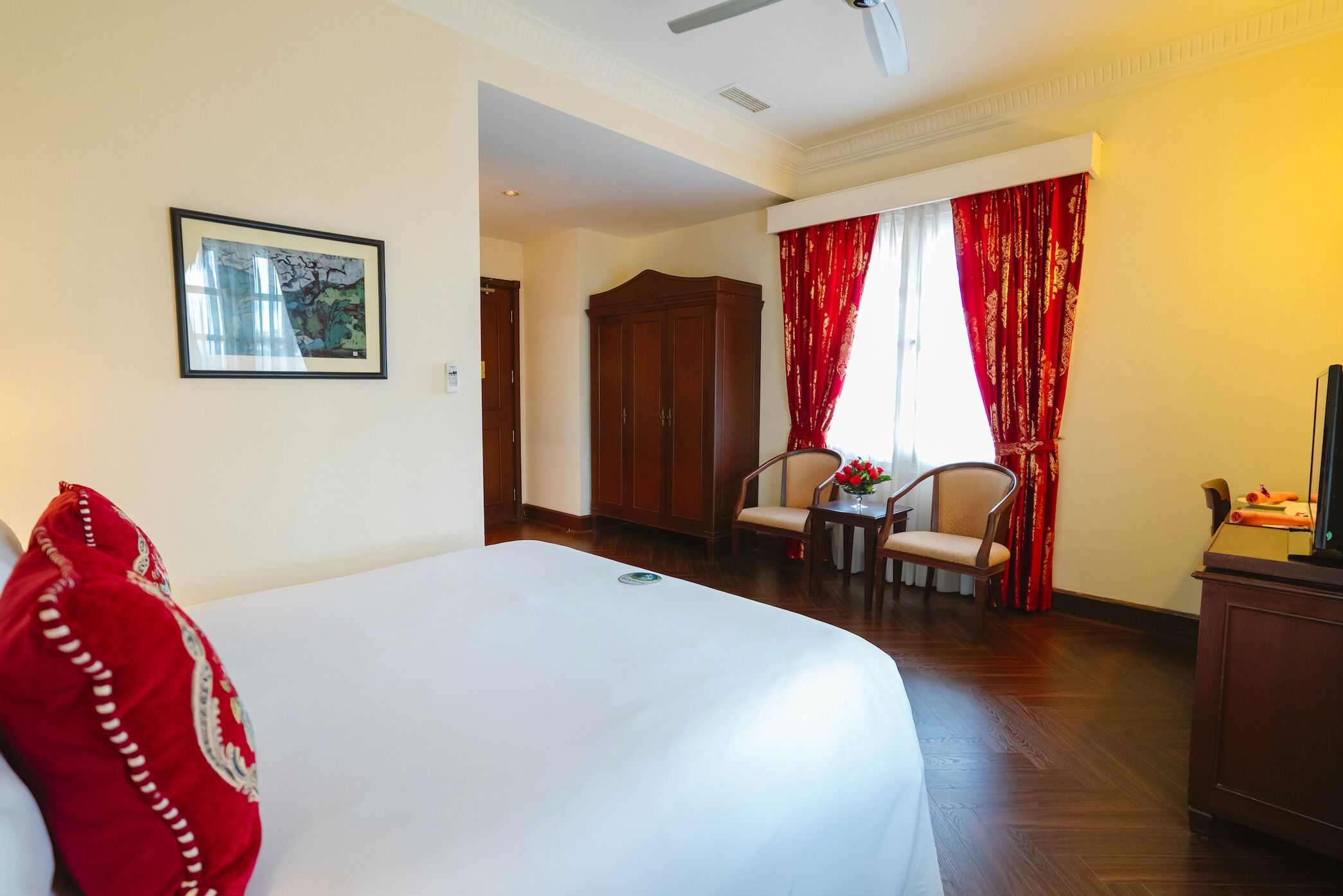 A hotel room with a bed, a chair, a small table, a wardrobe, and a window with red curtains.