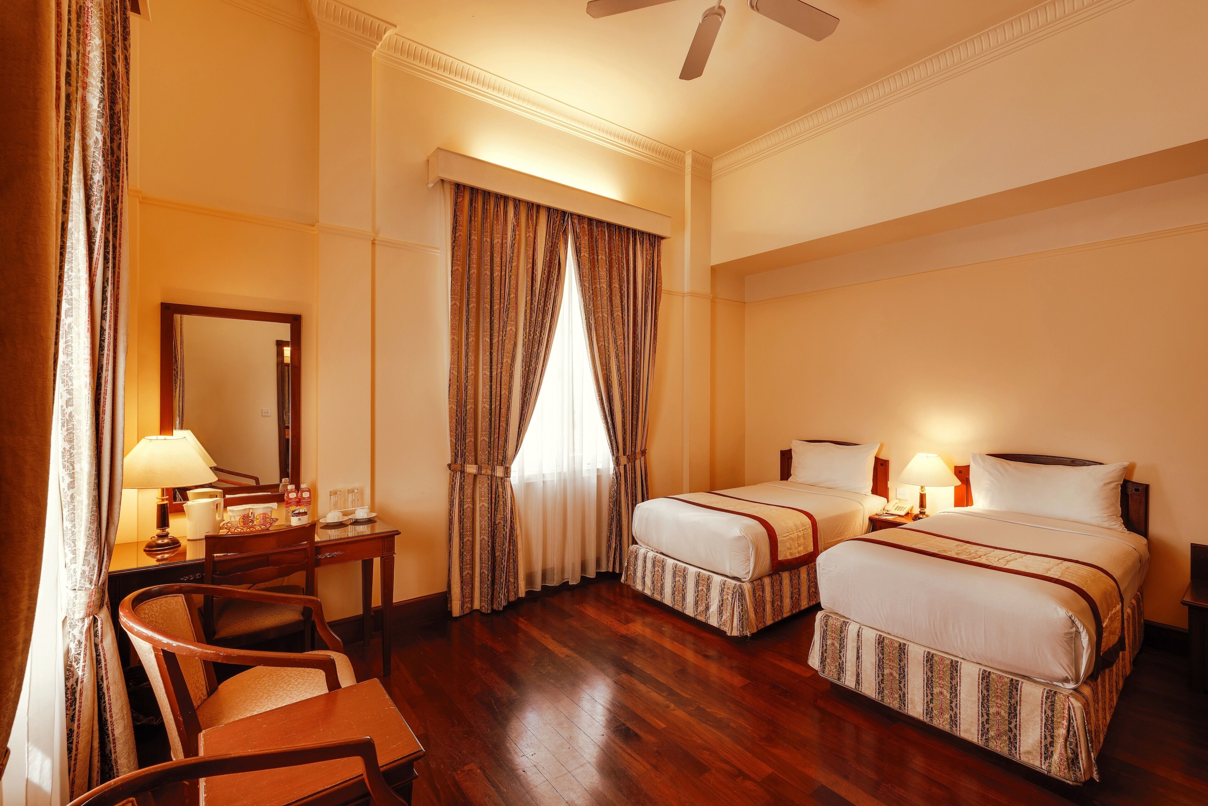 Ett hotellrum med två sängar, ett skrivbord med en lampa, en stol, ett fönster med gardiner och en takfläkt.