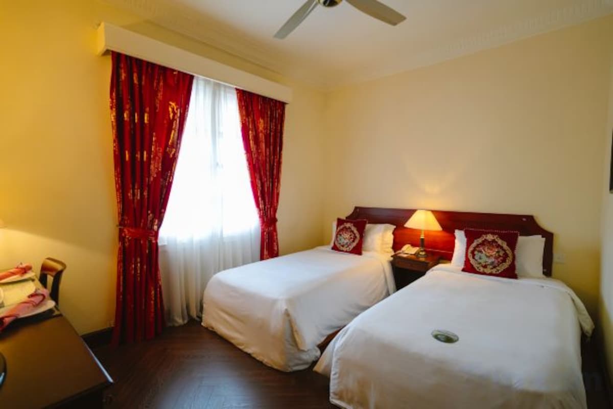 A hotel room with two beds, a desk, and a window with red curtains.