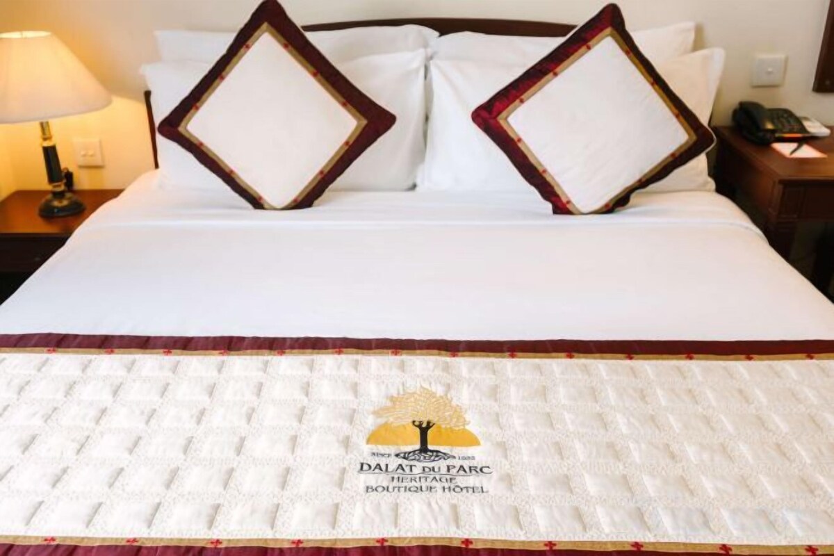 A hotel bed with a white bedspread and a logo, two decorative pillows, a nightstand with a lamp, and a phone.