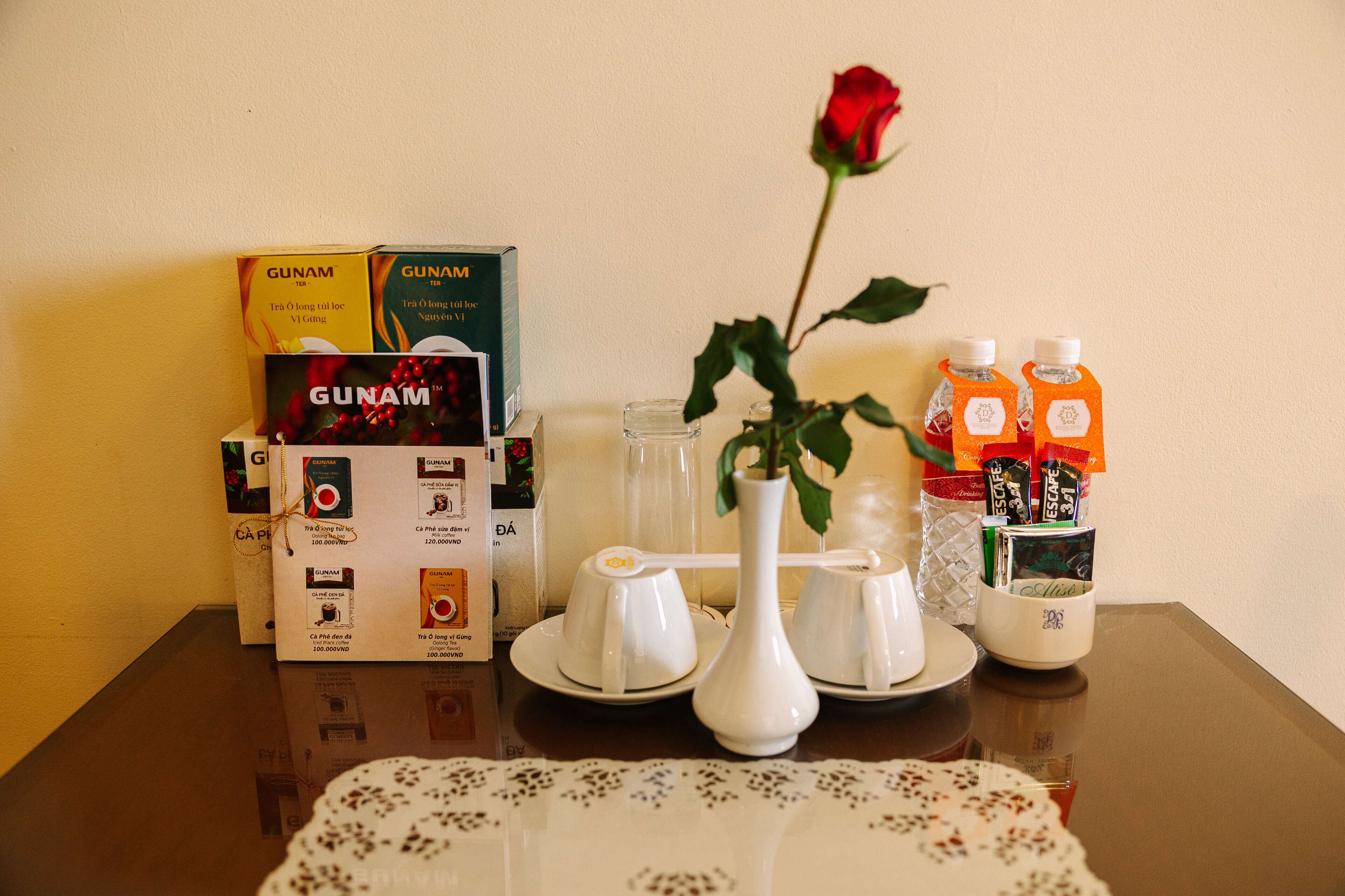 Una mesa con cajas de té GUNAM, un jarrón blanco con una rosa y dos botellas de jugo de naranja.