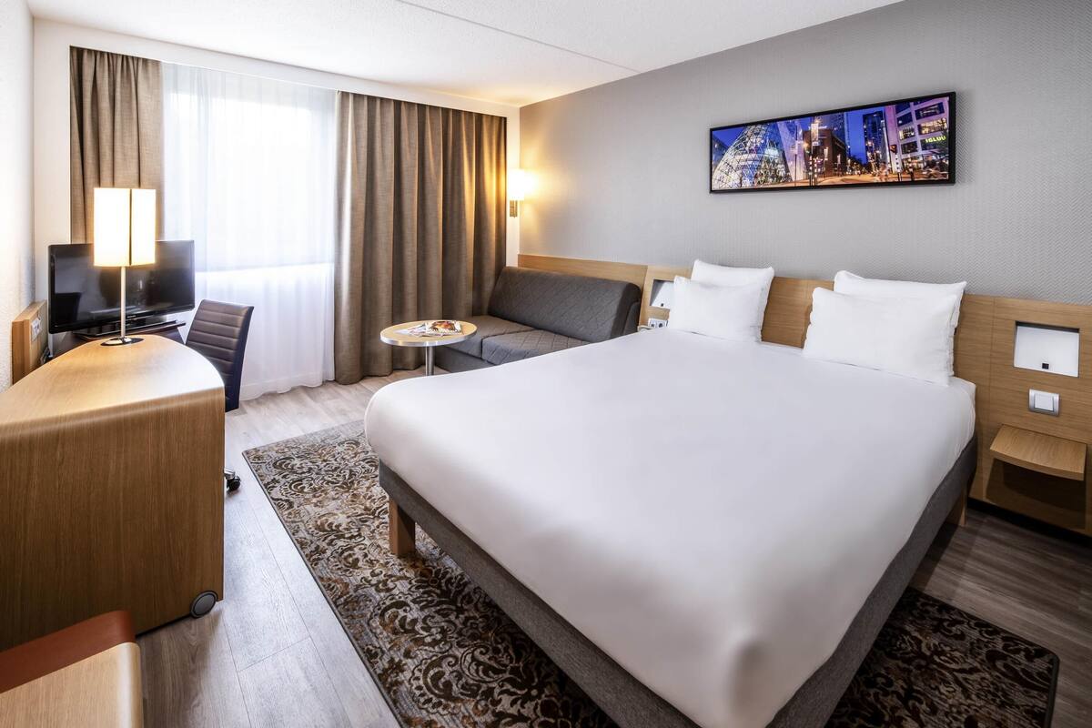 A hotel room with a large bed, a desk with a TV, a sofa, and a painting on the wall.