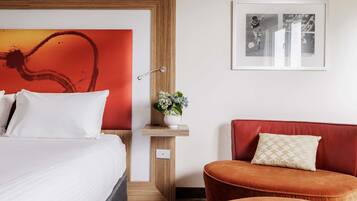Et hotelværelse med en seng, en rød polstret stol, et lille bord med en vase blomster og et indrammet billede på væggen.