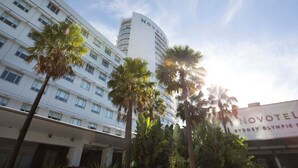 A hotel with a modern design, featuring a prominent 'NOVOTEL' sign, multiple floors, and a surrounding area with palm trees.