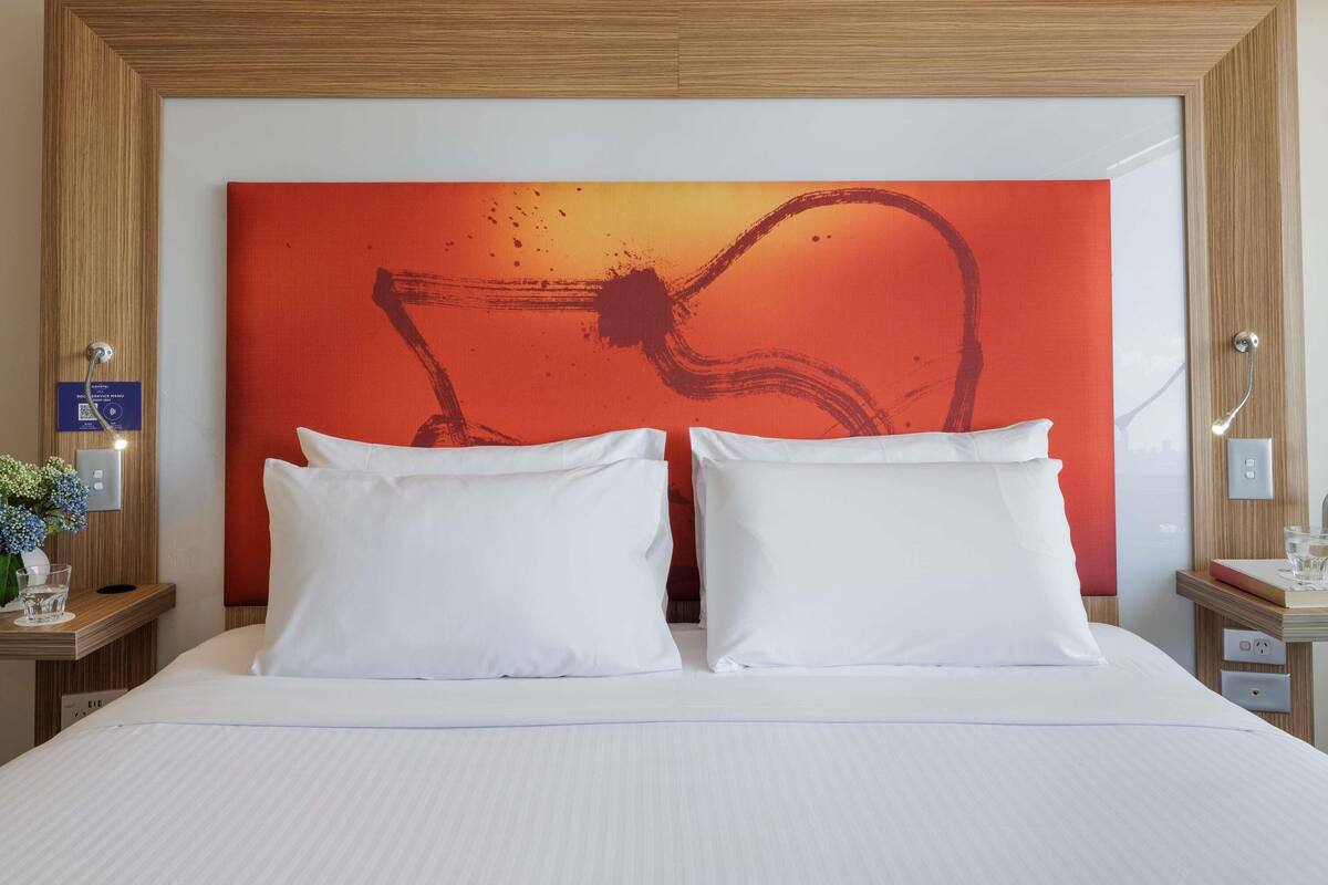 Habitación de hotel con una cama grande, dos almohadas blancas, un cabecero de madera y un cuadro abstracto rojo en la pared.