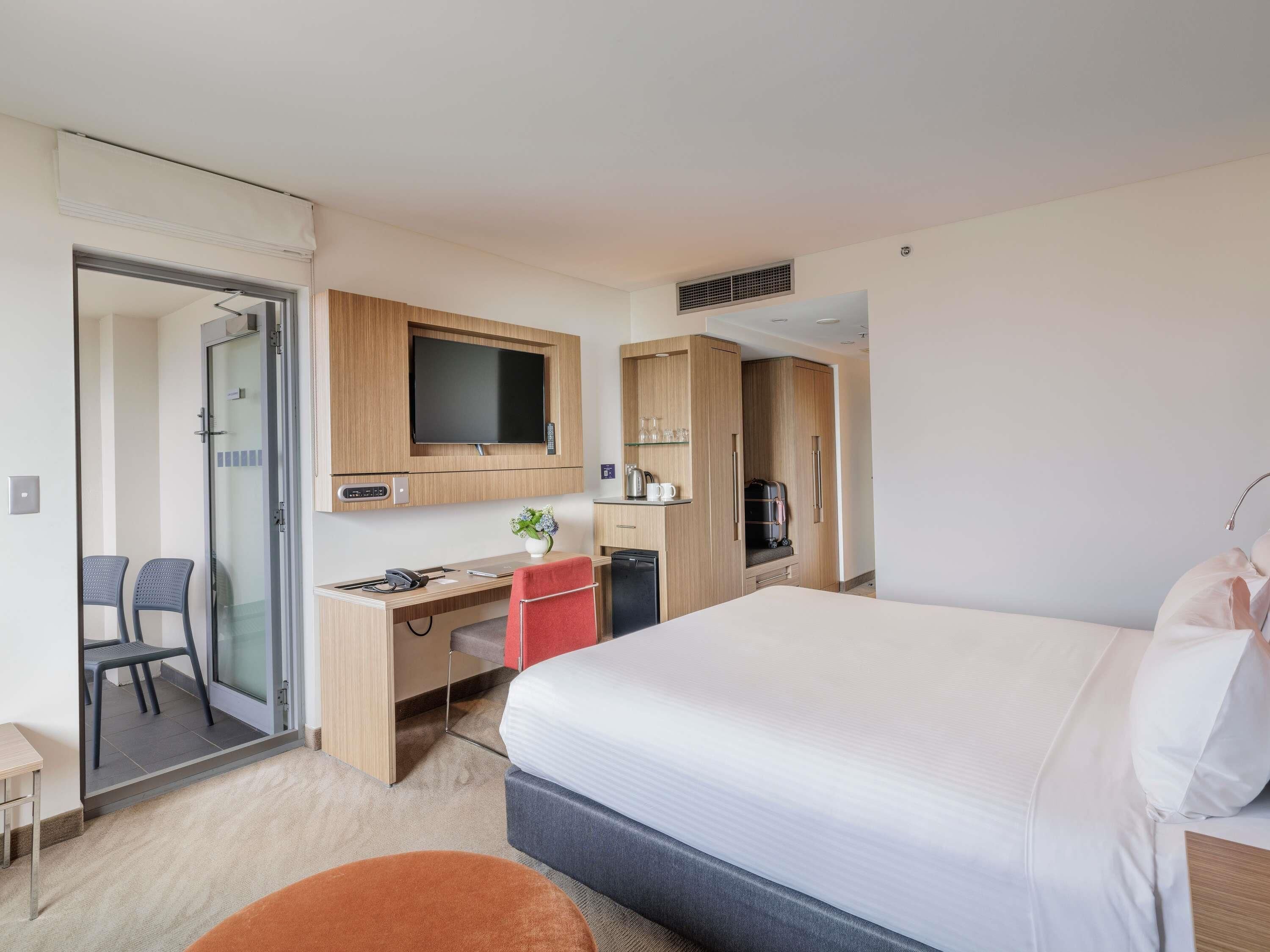 A hotel room with a bed, desk, chair, TV, wardrobe, and a glass door leading to a shower area.