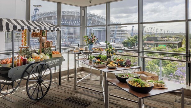 A dining area with a view of the Accor Stadium, featuring a food cart and various dishes on tables.