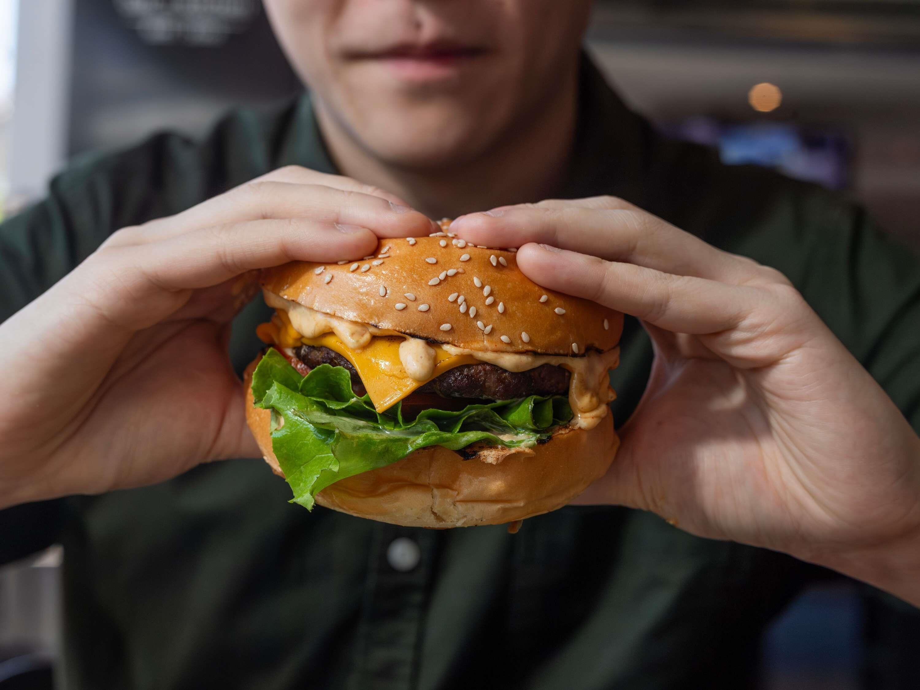 A person holding a burger with sesame seed bun, lettuce, cheese, and a beef patty.