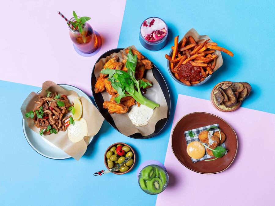 Variedad de platos que incluyen alitas, papas fritas y un postre, dispuestas sobre una mesa con fondo azul y rosa.
