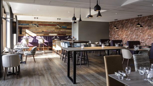 A modern restaurant with wooden flooring, black pendant lighting, and brick walls. Tables are set with white tablecloths and silverware.