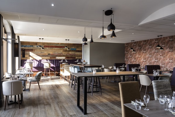 A modern restaurant with wooden flooring, black pendant lighting, and brick walls. Tables are set with white tablecloths and silverware.