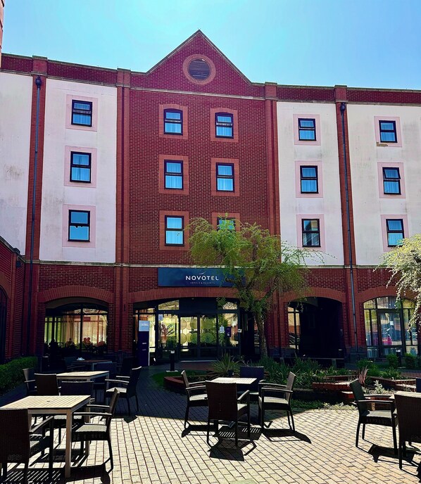 Un hôtel à la façade en briques, doté de nombreuses fenêtres et d’un espace extérieur aménagé avec des tables et des chaises.