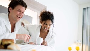 A couple in white bathrobes sitting on a bed in a hotel room, with breakfast items and two glasses of orange juice on the side table. - Novotel Ipswich (Ipswich)