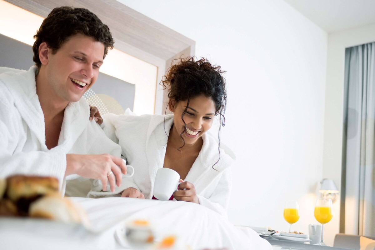 Una pareja con batas blancas sentada en una cama en una habitación de hotel, con alimentos para el desayuno y dos vasos de jugo de naranja sobre la mesita de noche.