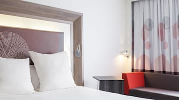 A hotel room with a bed, a nightstand, a sofa, a wall-mounted lamp, and a window with curtains.