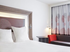 A hotel room with a bed, a nightstand, a sofa, a wall-mounted lamp, and a window with curtains. - Novotel Ipswich (Ipswich)