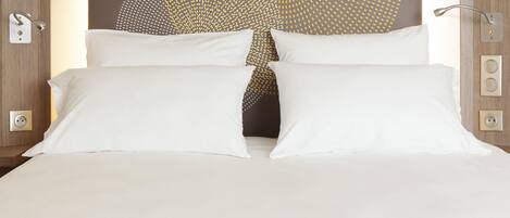 Una cama con ropa de cama blanca, un cabecero con diseño en la pared y una mesita de noche con una lámpara y un enchufe.