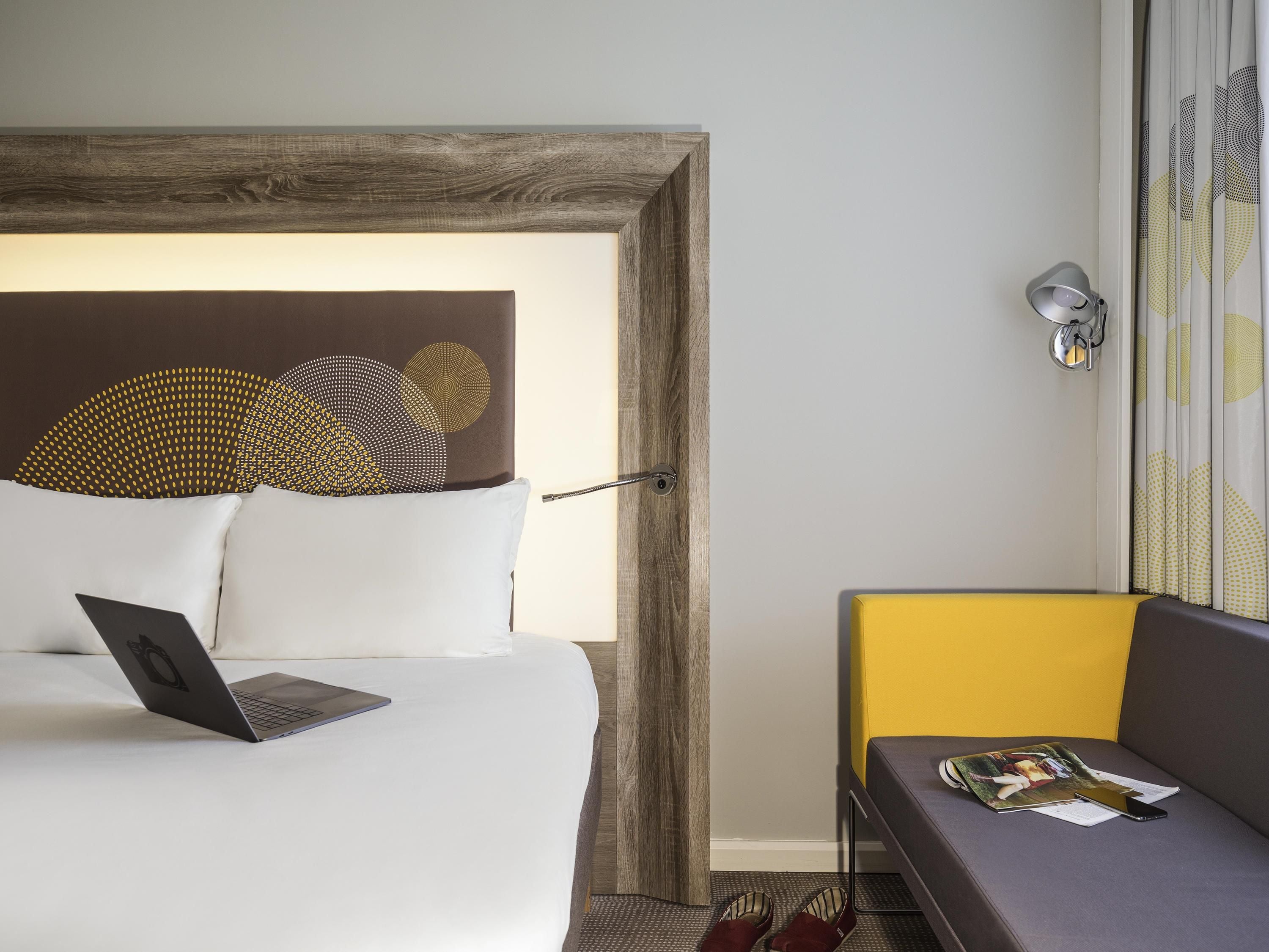 A hotel room with a bed, a laptop, a magazine, and a small table with slippers.