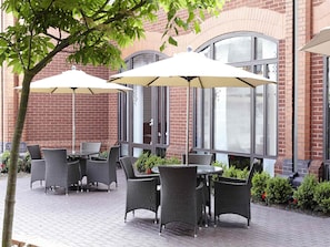 Une terrasse extérieure avec des meubles en osier et des parasols, un bâtiment en briques en arrière-plan et un arbre au premier plan.