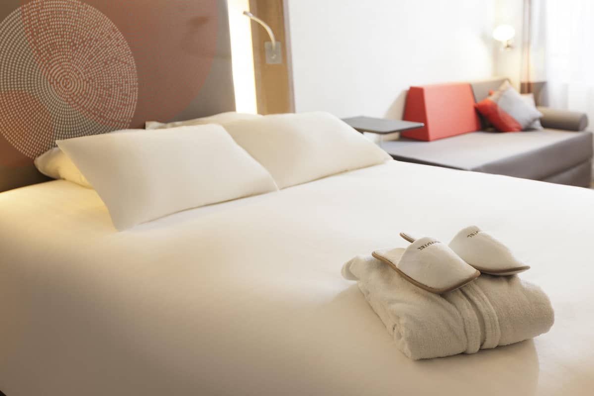 A neatly made bed with pillows, a bedside table with a lamp, and a red box on a sofa.