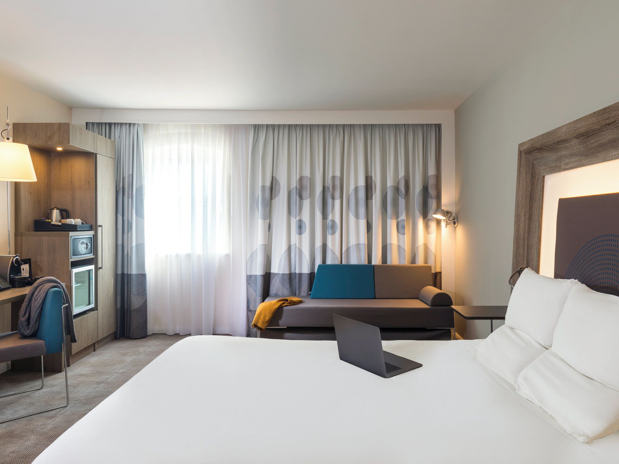 A hotel room with a bed, a desk with a laptop, a sofa, a chair, a lamp, and a window with curtains.