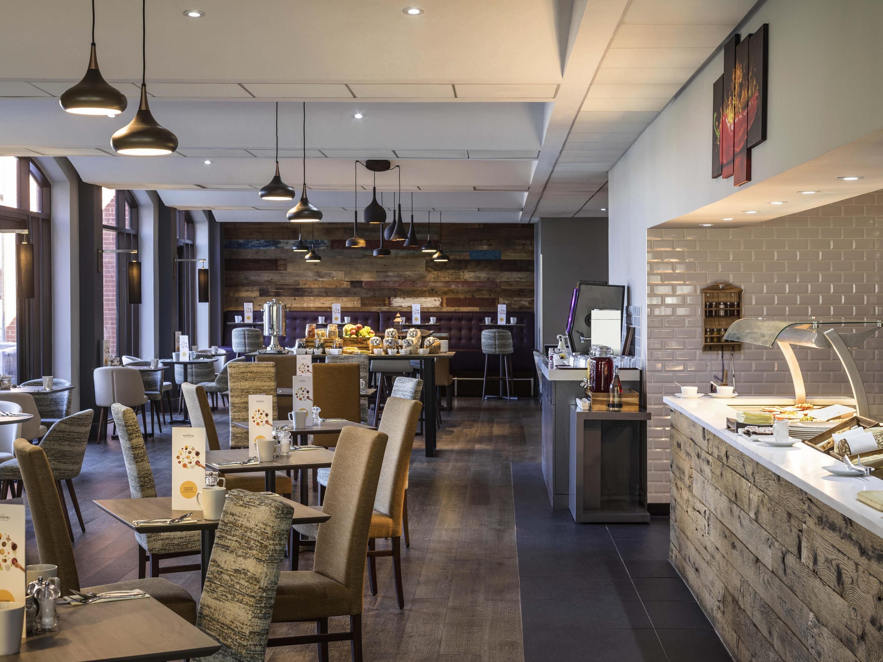 A modern restaurant with high ceilings, pendant lighting, and a brick accent wall. Tables are set with chairs, and a bar area is visible in the background.