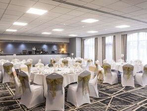 A conference room with round tables, white chairs with gold sashes, a bar area, and large windows.