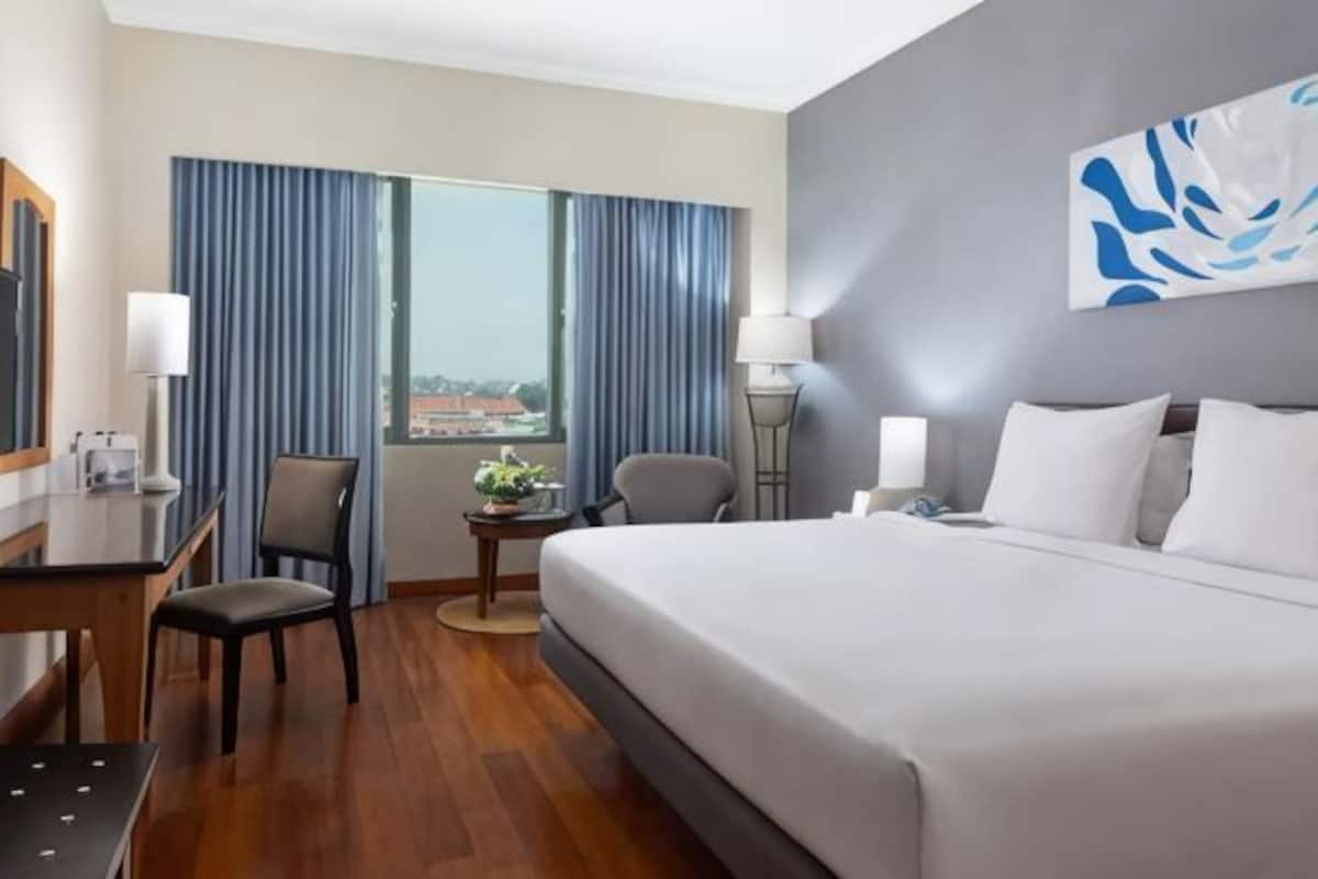 A hotel room with a bed, desk, chair, lamps, and a view through the window.
